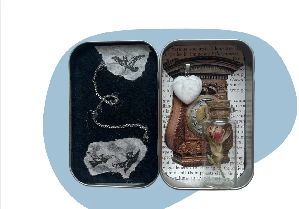 An opened tin containing a miniature assemblage of small found objects, its lid laid beside it, on a pale blue backdrop.