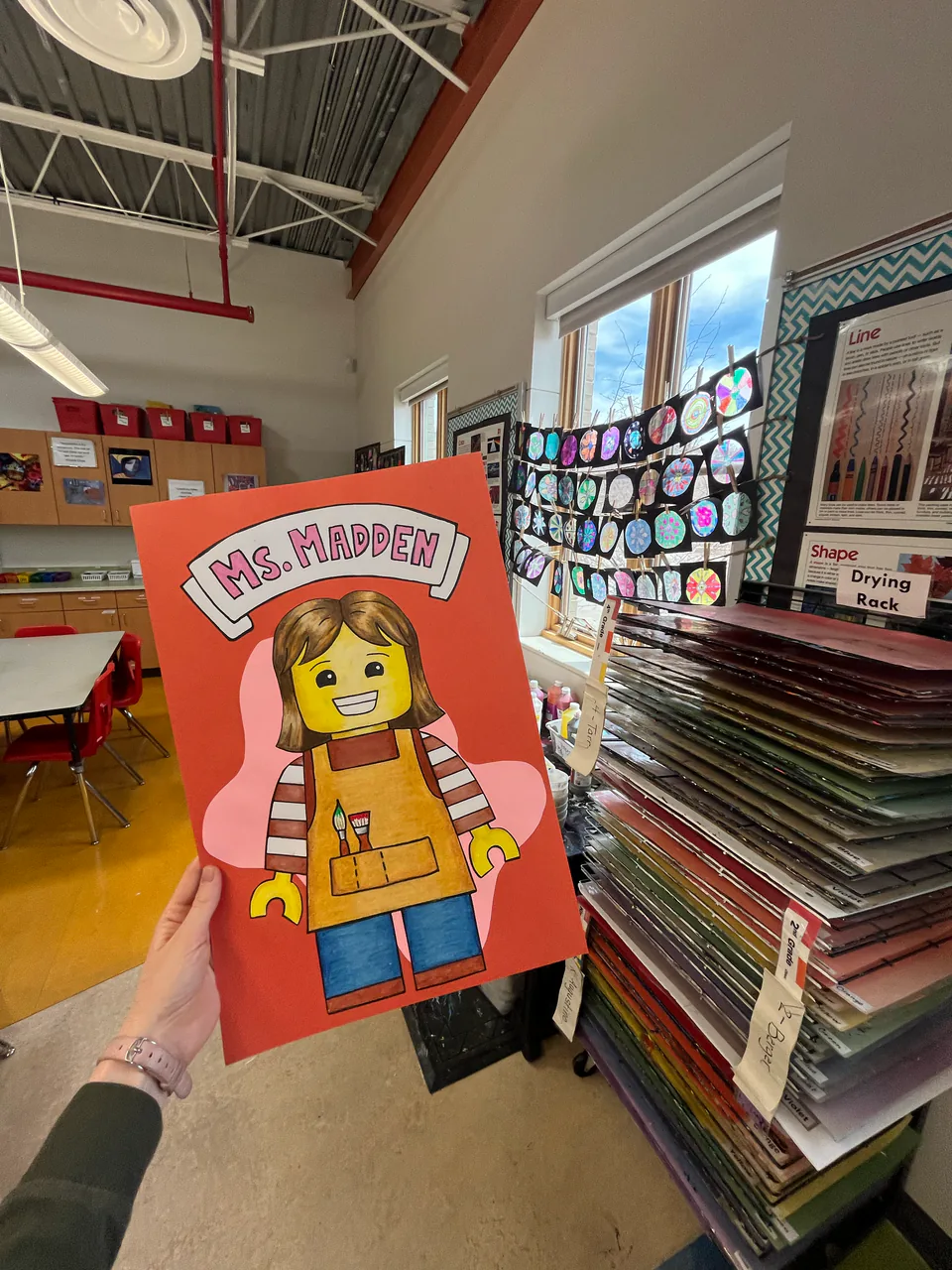 A hand holds up a child’s paper collage figure, built from strips of bright coloured paper, in an elementary art room.