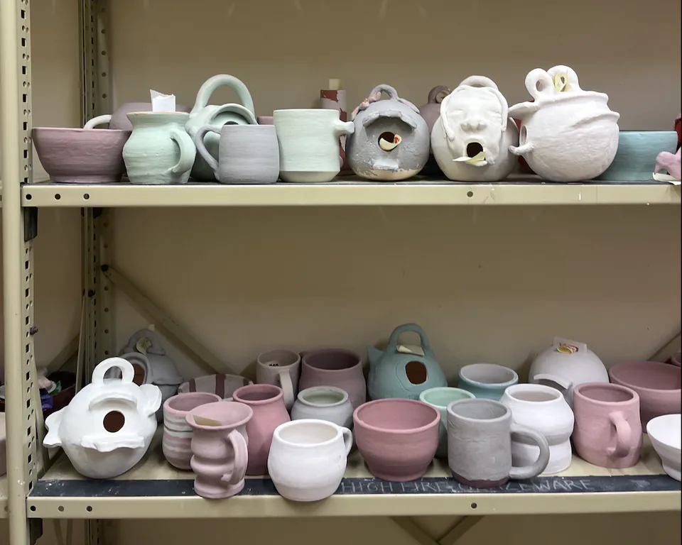 Shelves filled with student ceramic teapots and vessels in pale glazes.