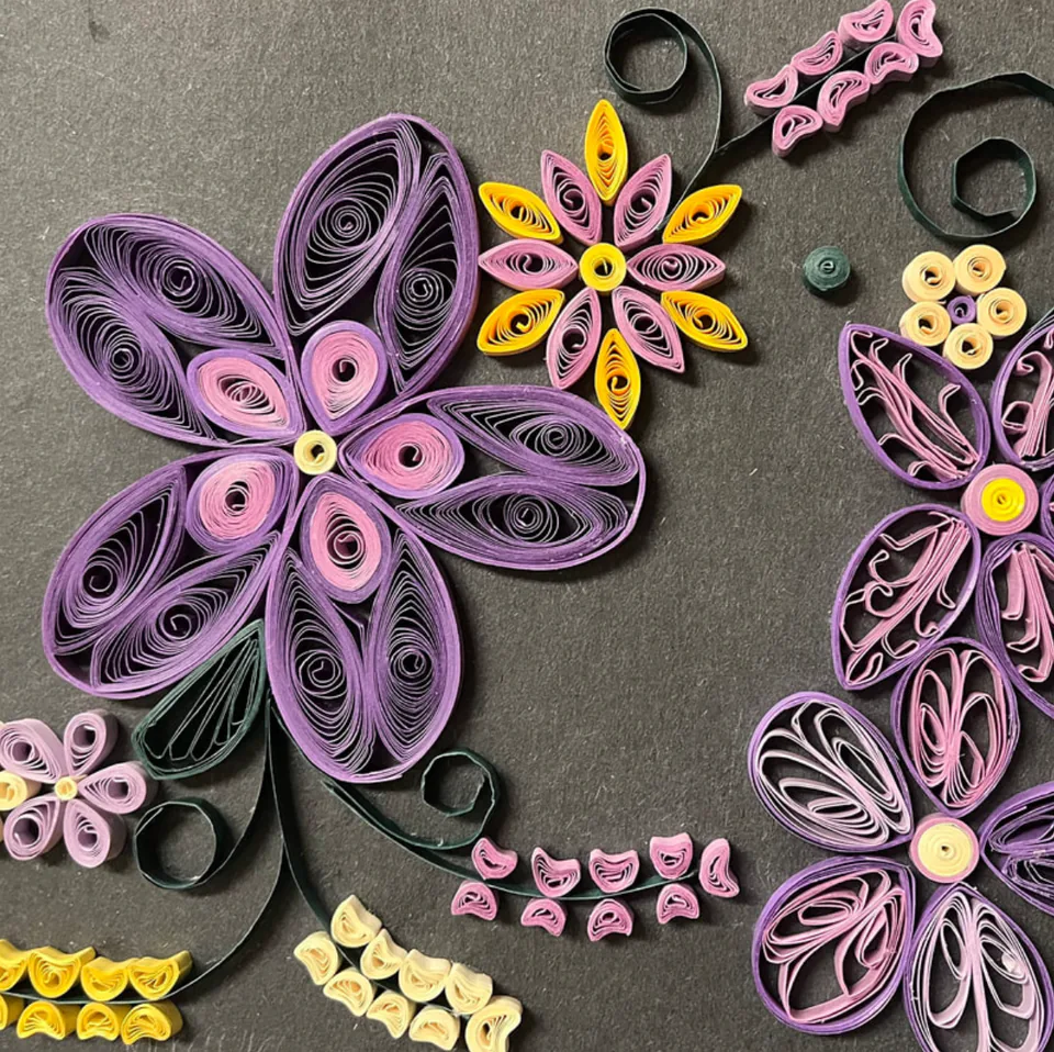 Paper quilling: coiled paper flowers in pink, purple and yellow arranged on a grey ground.