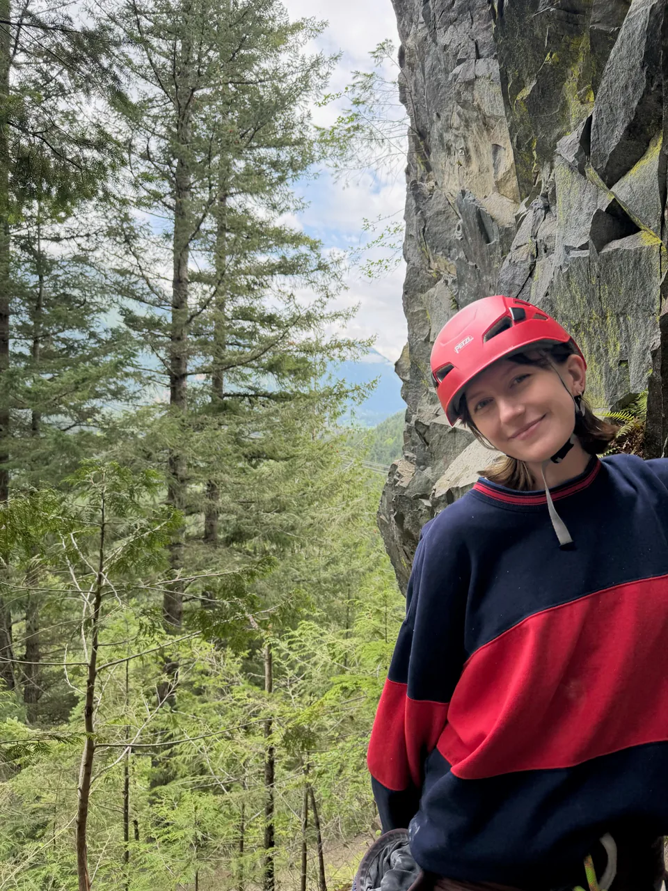 Sarah in a red climbing helmet, smiling, with pines and a rock face behind her.