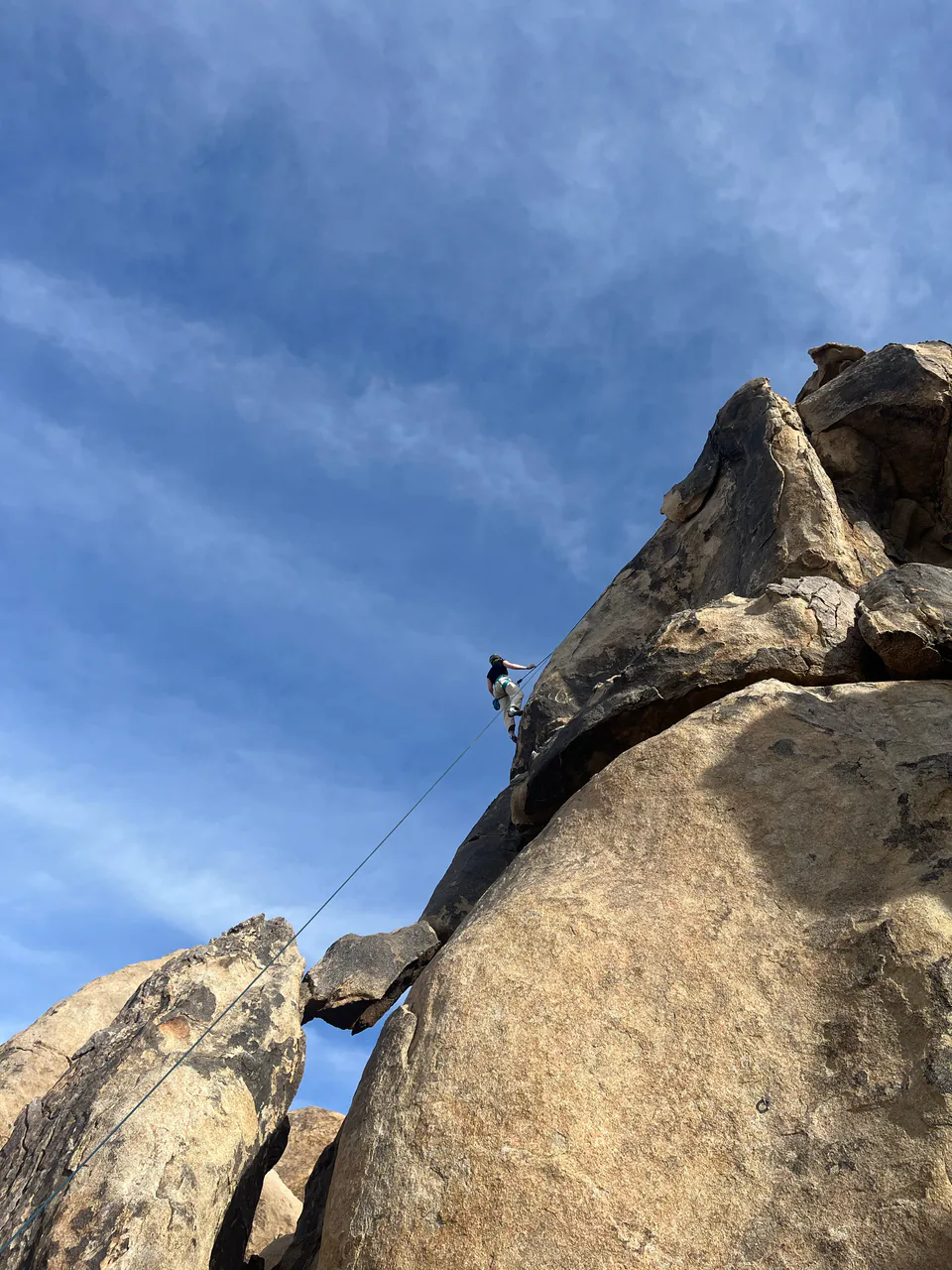 A climber high on a boulder, seen from below against a clear blue sky.