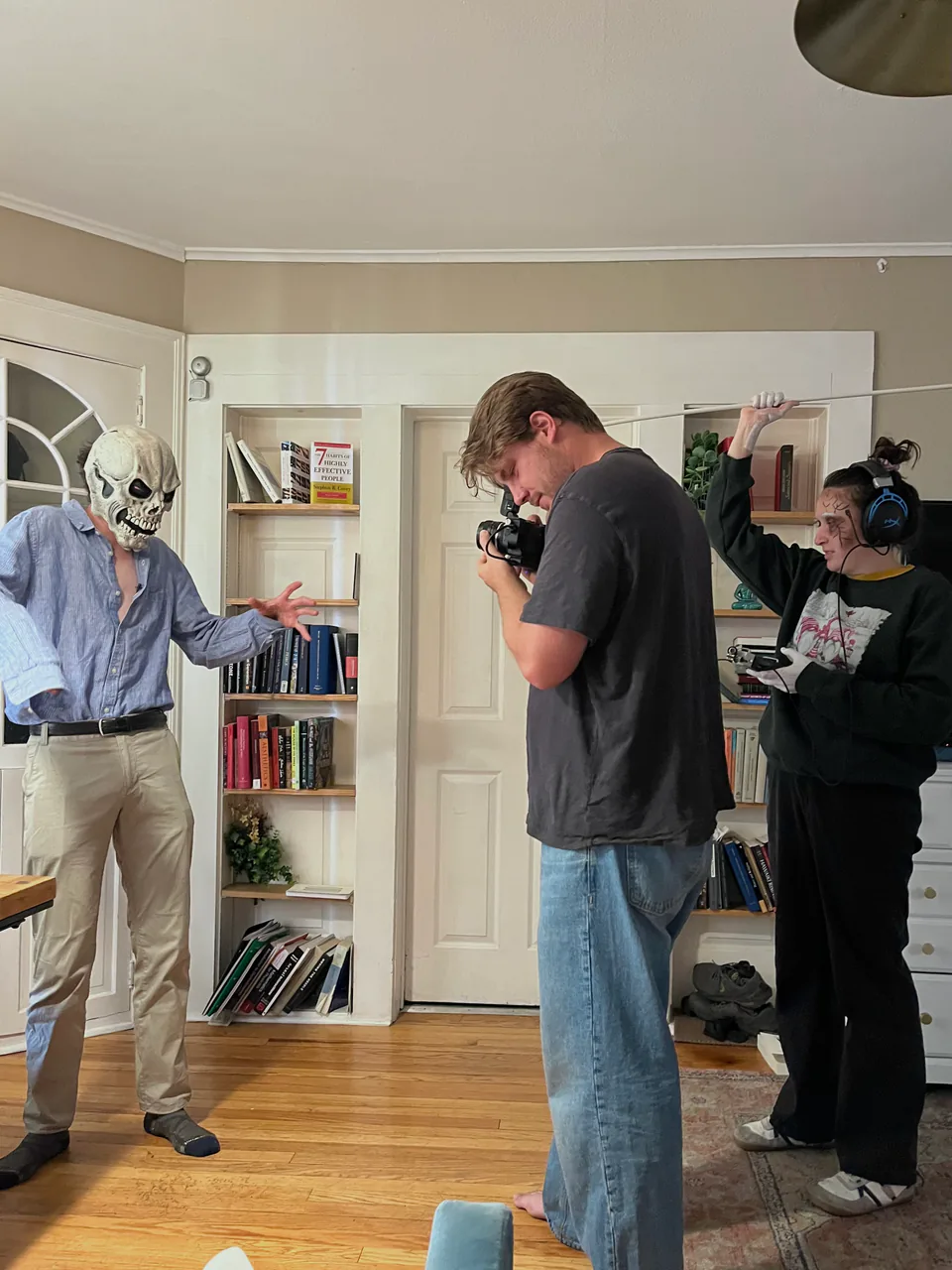 Three friends filming at home — one in a white mask, another holding a camera, in front of a bookshelf.