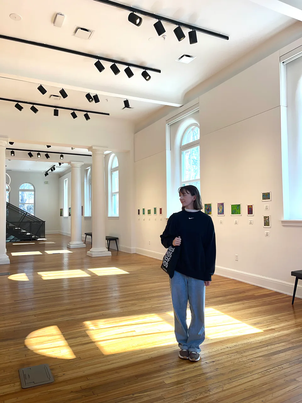 Sarah standing in a sunlit gallery with tall arched windows, a row of small paintings hung along the wall behind her.