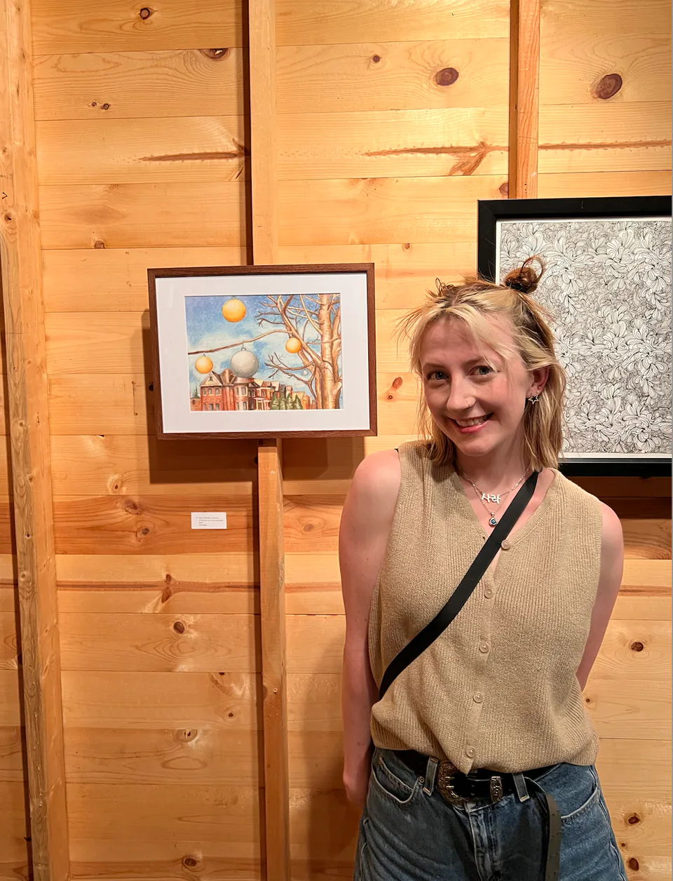 Sarah standing beside a small framed painting hung on a wood-panelled wall.
