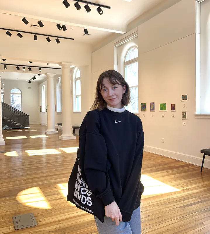 Sarah in a sunlit gallery, a canvas tote over one shoulder, with a row of small paintings hung on the wall behind her.